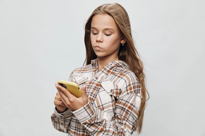 Girl with long hair wearing plaid shirt holding smartphone focused on screen for texting or social media communication on light background indoors with casual style and serious expression.