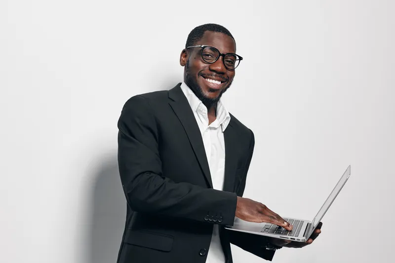African American man with dark skin wearing glasses and black suit smiling while holding laptop for business or technology concept on white background. Professional male with confident pose.