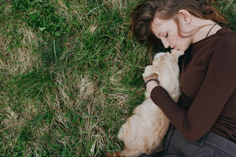 Young woman lying on grass gently cuddles a baby goat in a tender moment of connection and relaxation outdoors during a casual lifestyle scene.