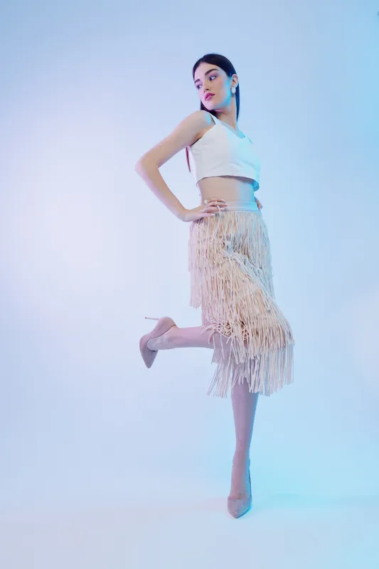 Young woman beauty fashion model posing in a white crop top and fringed skirt. Studio shot with soft lighting on a gradient blue background. Concept for style, elegance, and confidence.