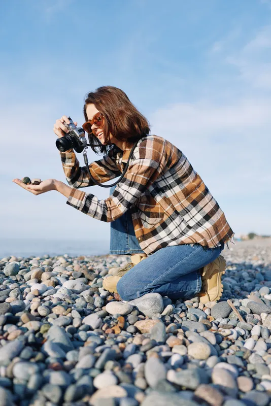 Woman photographer crouching on a pebble beach, capturing a shot with a camera in natural light. Plaid shirt, jeans and boots create a casual outdoor portrait on a rocky coast.