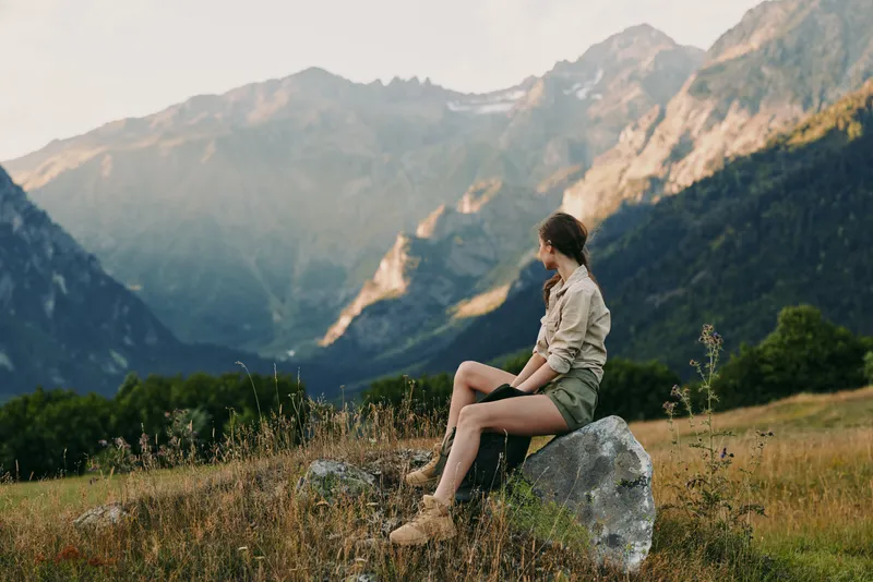 Young woman sits on a rock in a meadow with a distant mountain panorama, enjoying peaceful solitude in nature and feeling calm and free.