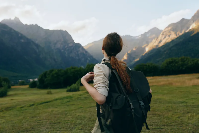 Woman with a backpack gazes over a mountain valley, exploring nature and outdoor life. Bright sky, green fields, distant peaks create a calm, adventurous mood for travel lifestyle.