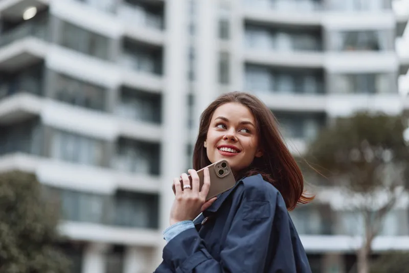 Young woman with long brunette hair wears a denim jacket and smiles while talking on a smartphone outdoors against a modern apartment building in bright daylight