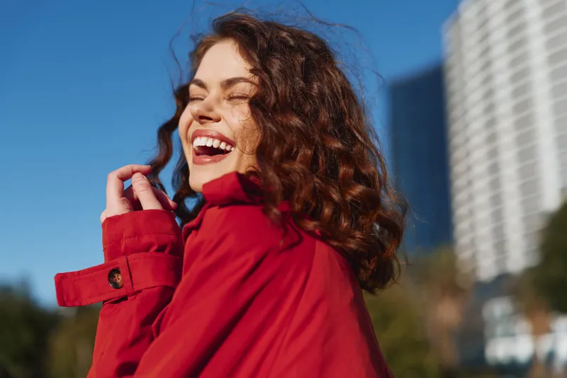 Young woman with curly hair in a red coat laughs on a sunlit urban street, capturing a carefree candid moment against modern buildings and a bright sky.