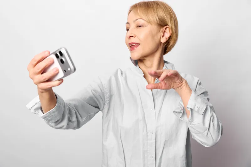 Smiling elderly woman holding a smartphone for a selfie, portrait on a light gray studio background, hand under chin, concept of mobile communication, technology, aging