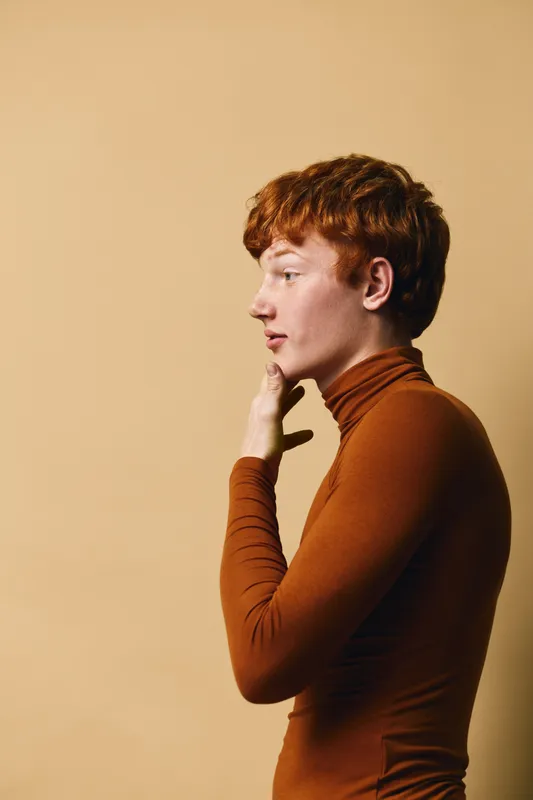 Young man with red hair wearing brown turtleneck sweater touching chin in thoughtful pose against beige background. Side view portrait with soft light on face and neck.