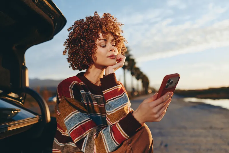 Woman lifestyle outdoors, curly hair, striped sweater, standing by open car at sunset, holding smartphone, relaxed face, warm breeze, scenic road and sky background soft light