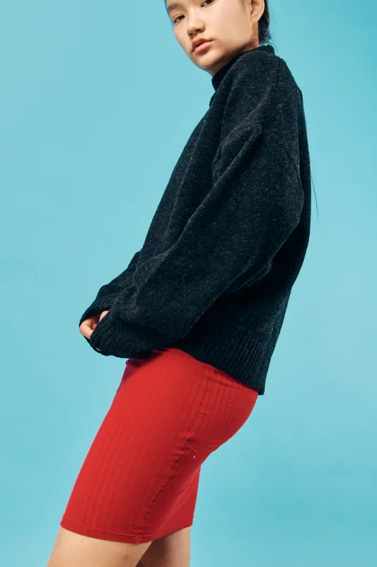 Young woman wearing black sweater with red skirt standing against light blue background. Casual outfit, fashion style, and confident pose for clothing or model concept in studio.