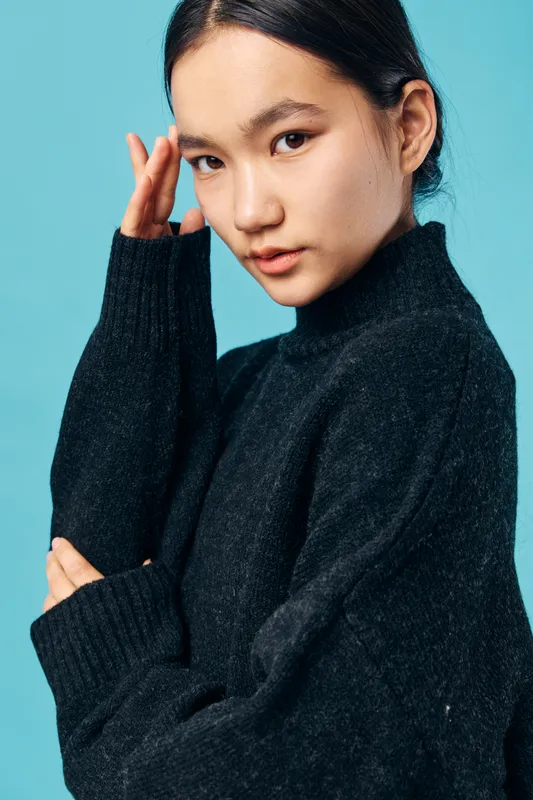 Young woman in dark sweater posing with hand near head against blue background for portrait photography, fashion, and casual style with thoughtful facial expression and natural lighting.