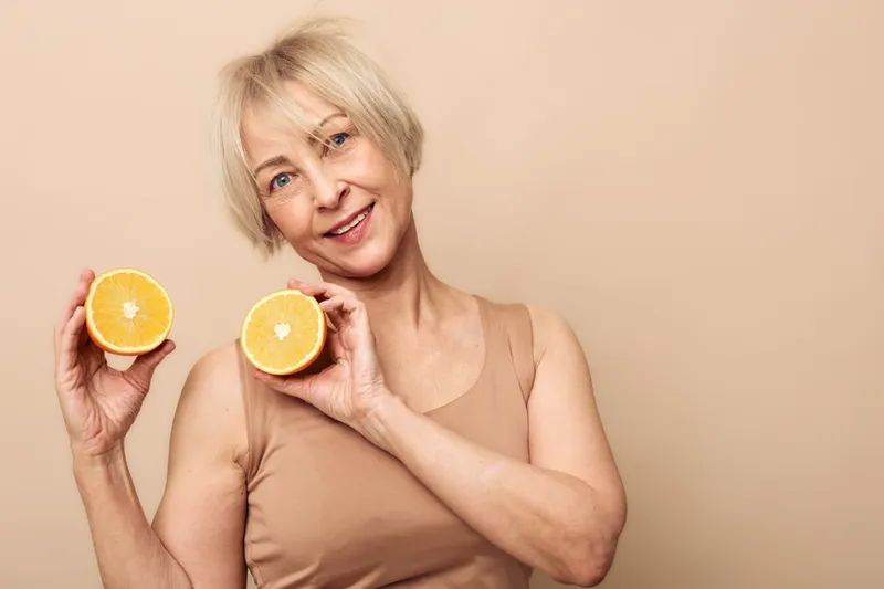 Smiling mature woman with short blonde hair holds two halves of fresh orange fruit, standing against beige background. Concept for healthy lifestyle, wellness, and natural vitamins.