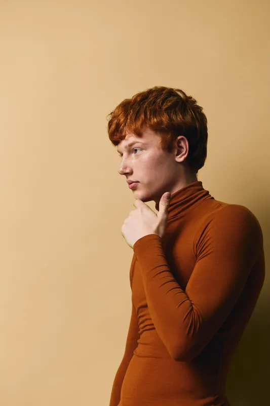 Young man with red hair wearing brown turtleneck sweater poses thoughtfully against beige background. Side profile, casual style, isolated portrait with modern fashion and contemplative mood.