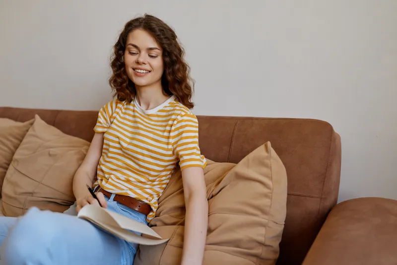 A woman sits on a sofa at home, smiling softly while writing in a notebook. A casual lifestyle scene in a cozy room with cushions, showcasing everyday creativity and focus.