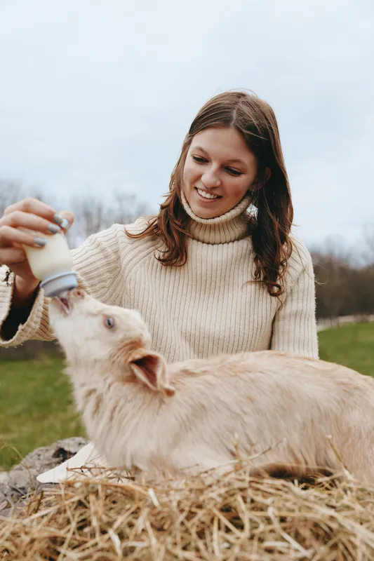 A woman bottle-feeds a goat kid outdoors in a rural setting, smiling as the animal drinks from the bottle, capturing care, bonding, and farm life in a natural lifestyle scene.