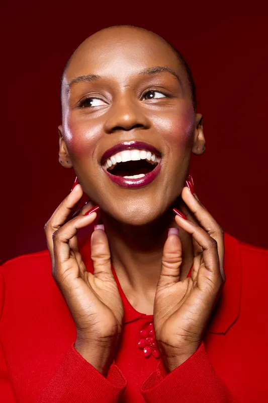 Happy African woman with dark skin and bold makeup smiles brightly wearing a red outfit. Close-up portrait on red background. Concept of beauty, fashion, and confidence.