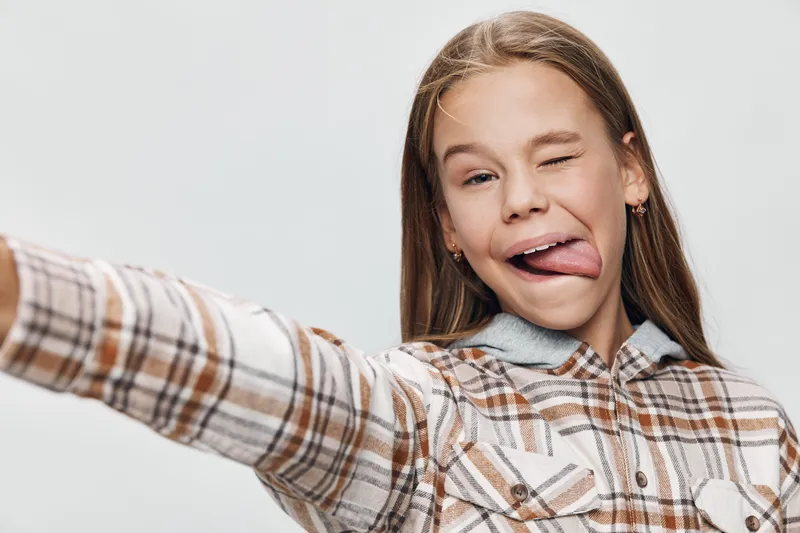 Girl taking selfie with tongue out and wink, casual plaid shirt, long hair and playful expression, isolated on white background for lifestyle and fun moments.
