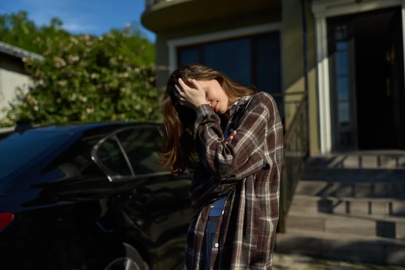 Woman outdoors in a plaid cardigan, casual lifestyle moment lit by sunlight, wind in her hair, relaxed pose, natural colors, warm background standing by a car, soft lighting, gentle breeze