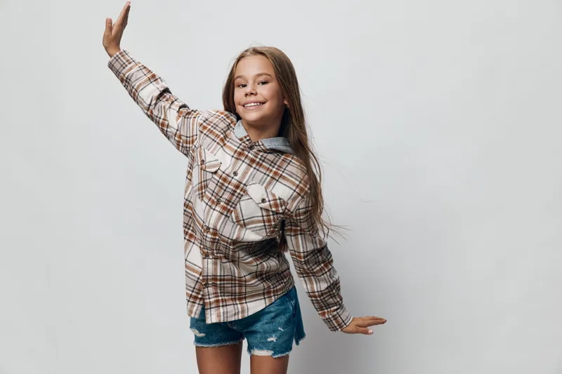 Girl smiling and dancing with raised arm wearing plaid shirt and denim shorts on white background. Happy child posing with casual outfit for lifestyle and fun activity.