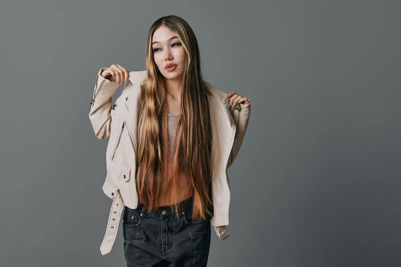 Young woman with long braided hair posing in casual outfit with light jacket and dark jeans against gray background in studio. Fashion, beauty and style concept.