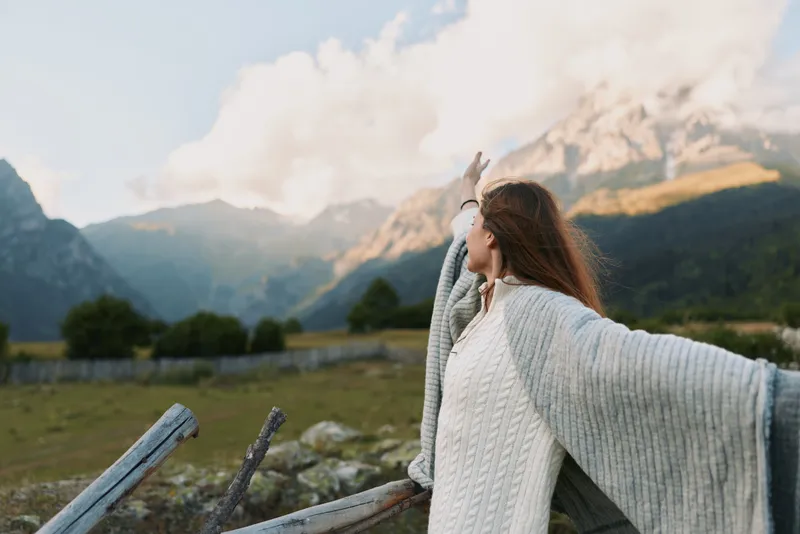 Woman with long hair wearing a white sweater and gray shawl stands outdoors with arms outstretched enjoying mountain landscape and fresh air in a peaceful nature setting.