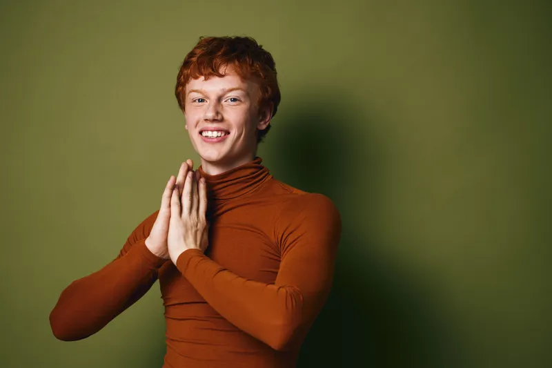 Young man with red hair wearing orange turtleneck stands with hands together smiling against green background. Portrait, casual, confident, isolated, studio lighting, happy mood.