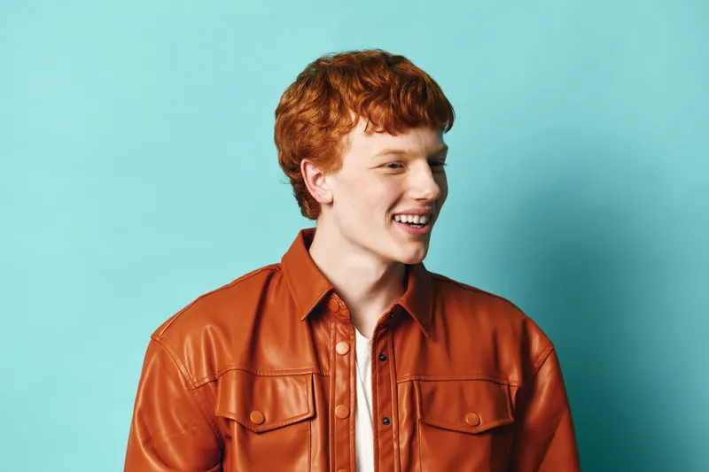 Young man with red hair wearing orange jacket smiling and looking to side on blue background. Casual portrait with natural expression and stylish clothing for fashion or lifestyle.