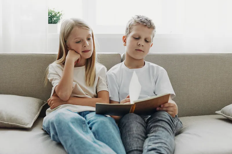 Two focused children sitting on a couch reading a book together in a bright living room. Concept of childhood learning, concentration, and quiet study time.