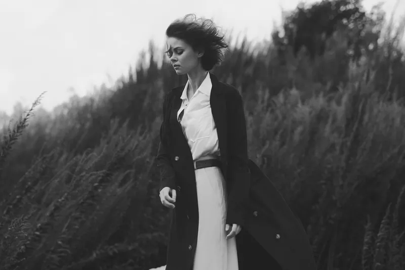 Monochrome image of a woman in a long coat walking through tall grasses, wind in her hair, moody fashion with outdoor cinematic vibe, timeless style and dramatic lighting