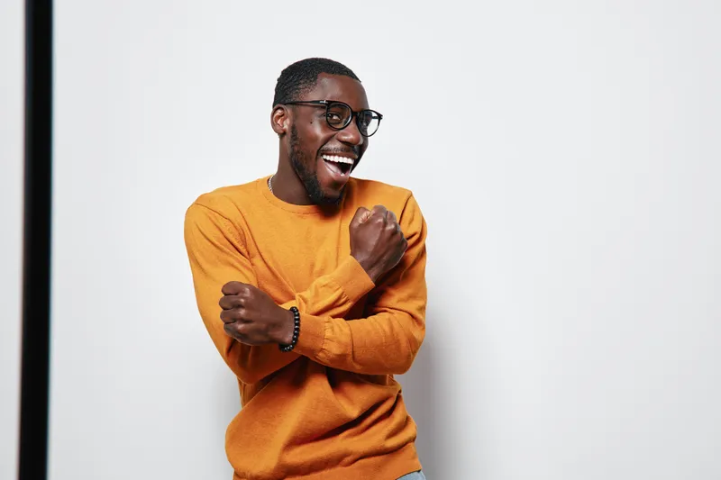 African American man with dark skin wearing glasses and orange sweater smiling and celebrating with crossed arms on white background in studio. Happy male portrait.