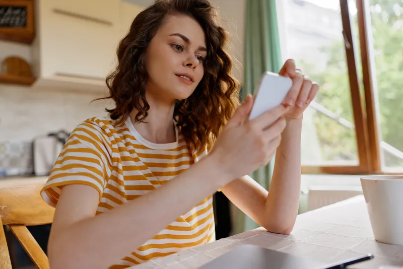 Young woman sits at a bright kitchen table, looking at her phone with a relaxed, curious expression and warm natural light.