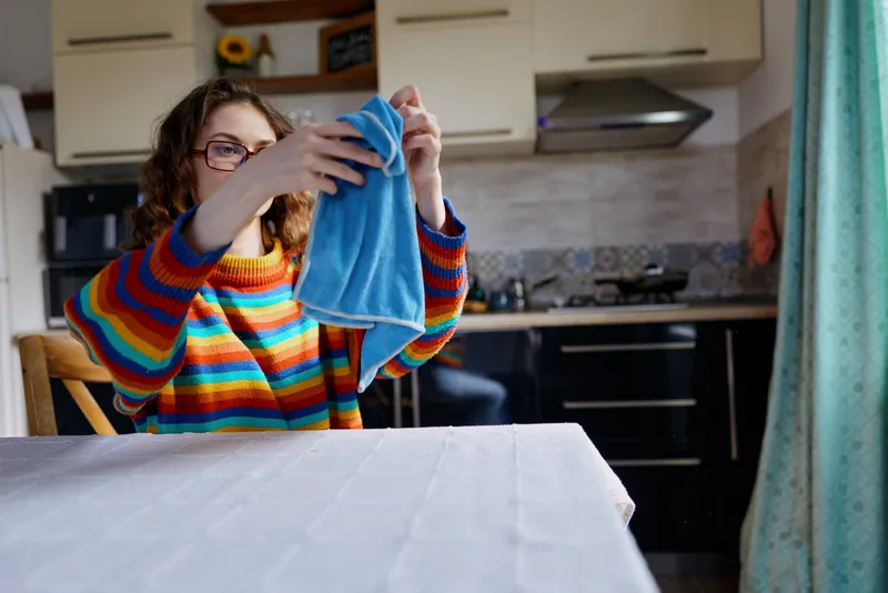 Bright kitchen scene shows a young woman with glasses drying a blue cloth over a table while wearing a colorful striped sweater and smiling in a cozy setting today.
