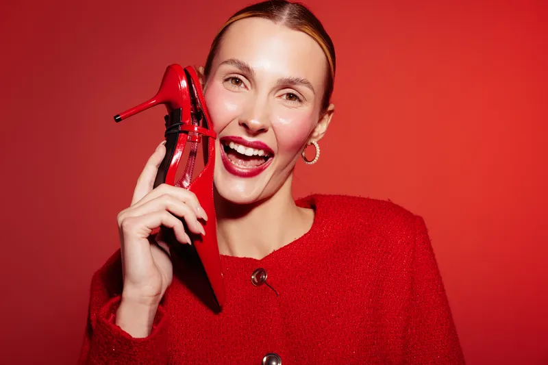Smiling woman in red sweater holds red high heel shoe near face on red background. Concept of fashion, style, happiness, and vibrant color contrast in studio portrait.