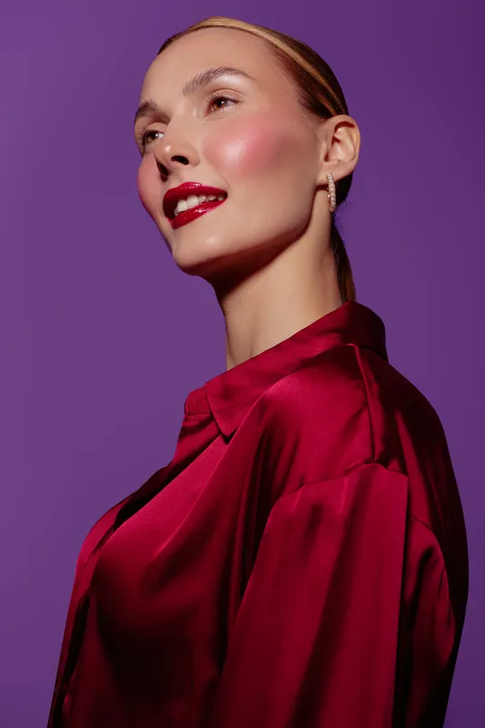 Portrait of a confident woman with slicked back hair wearing a red satin blouse against purple background. Makeup features glossy red lips and glowing skin. Fashion and beauty concept.