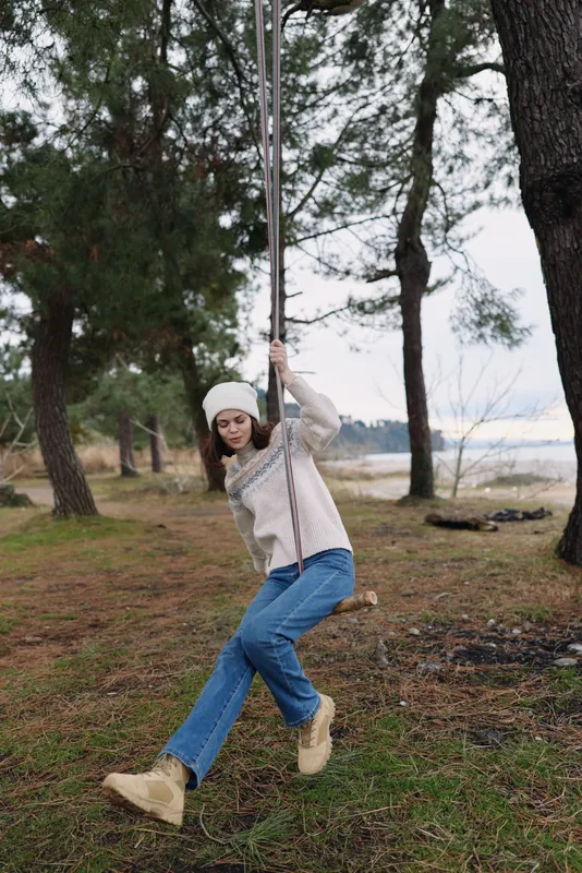 Outdoor woman swinging on a rope swing among trees in a forest, casual sweater and jeans, playful moment in nature and leisure in a calm outdoor setting