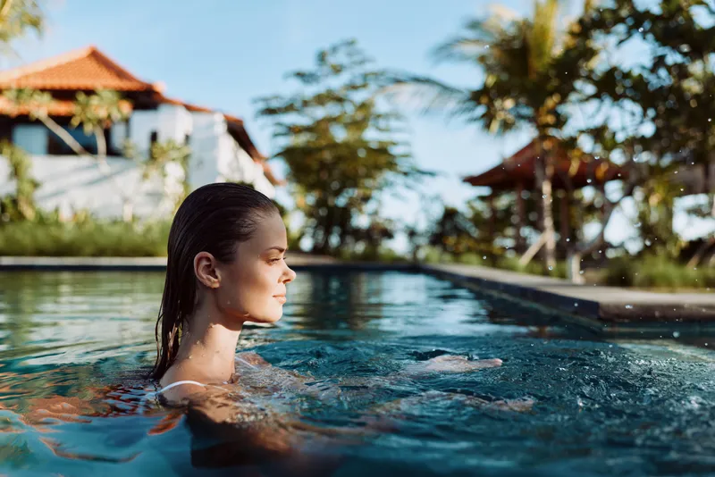 A woman rests in a clear pool with head above water, gazing toward the sunny resort. The scene conveys relaxation, escape, and personal wellbeing in a casual lifestyle setting.