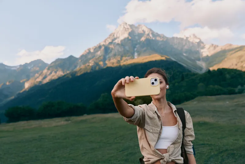 A woman in casual attire takes a selfie outdoors in a green field, with mountains and blue sky behind, highlighting lifestyle moments in nature and travel.