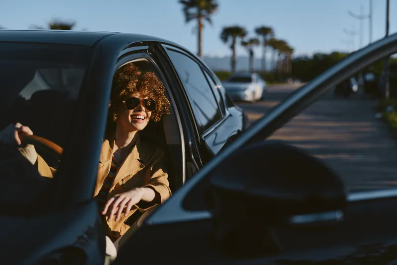 A stylish woman sits in a car, smiling through the window in warm light. A casual lifestyle moment with retro film color and sunny street vibe.