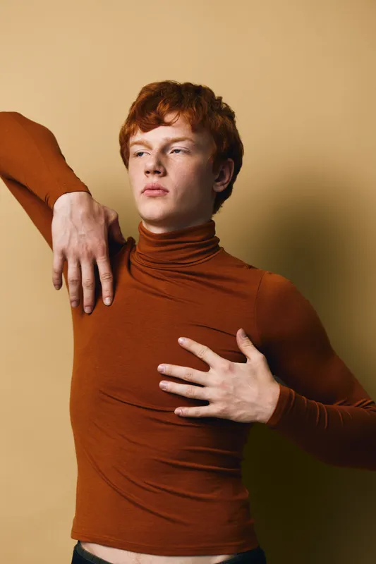 Redhead man in brown turtleneck stretching with arms raised on beige background. Male model posing with expressive face and casual clothing in studio portrait session.