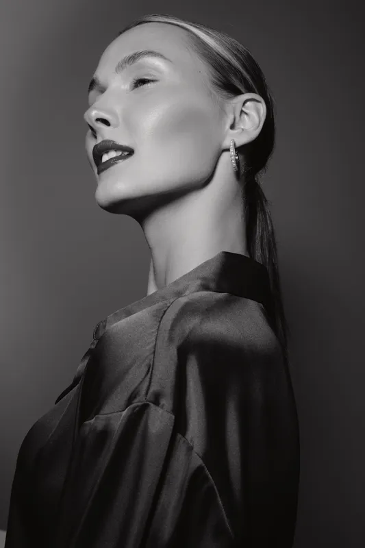 Elegant white woman with fair skin and sleek hair poses in profile wearing shiny blouse and hoop earrings in black and white studio portrait.
