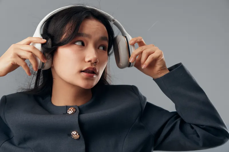 Young woman wearing headphones adjusting them with hands looking sideways with thoughtful expression in studio. Asian female dressed in black jacket with buttons on gray background.