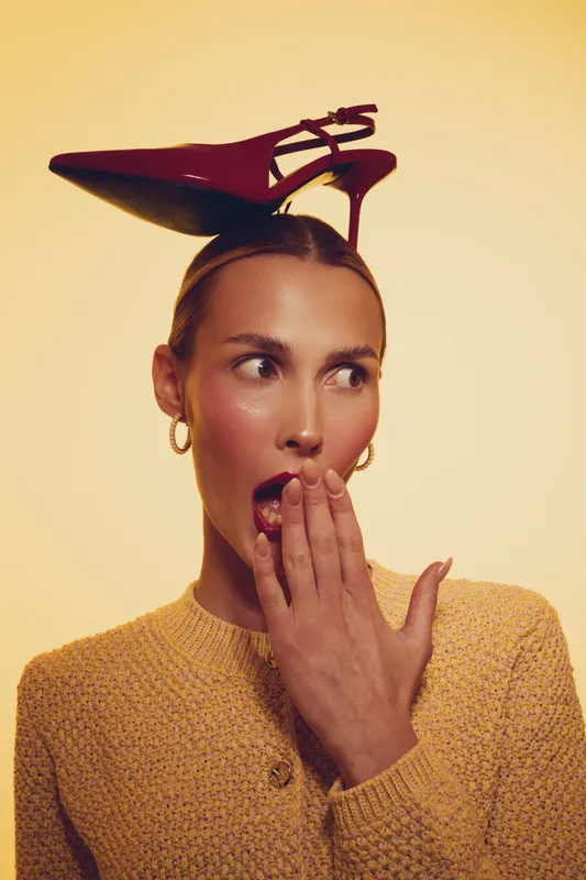 Surprised woman with slicked hair holds red high heel shoe on her head, wearing yellow sweater and gold hoop earrings. Concept for fashion, style, and playful attitude in studio lighting.