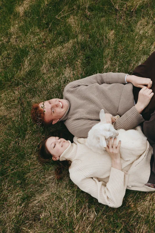 couple lifestyle outdoor grass scene featuring a relaxed duo lying on the lawn with a curious goat kid, cozy sweaters, warm smiles, peaceful moment, natural light, candid family vibe