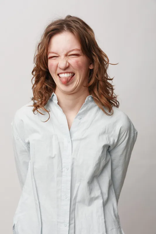 Young woman with curly hair wearing white shirt makes funny face sticking out tongue. Studio portrait on light background. Concept of humor, playfulness, and casual lifestyle.