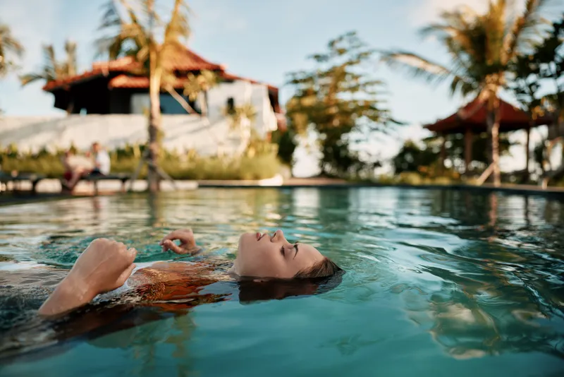 Young woman floating calmly on water in an outdoor pool surrounded by tropical palm trees and a sunny sky. Concept of relaxation, lifestyle, and summer vacation.