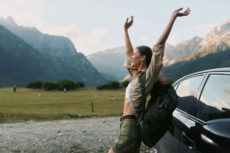 A woman with a backpack stands by a car in a mountain valley, raising her arms in celebration, enjoying open space and a sense of freedom.