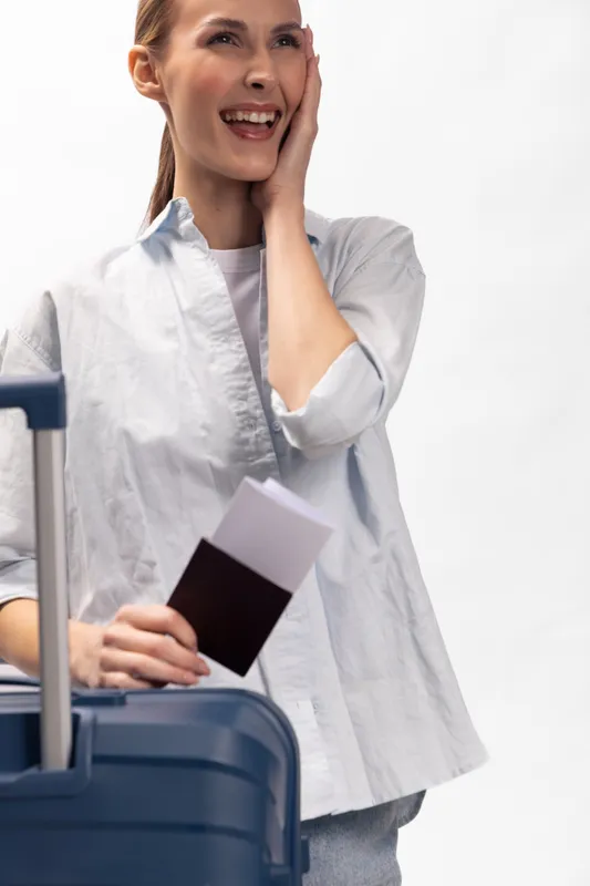 Happy young woman holding passport and airplane ticket stands near blue suitcase, smiling and touching face. Concept for travel, vacation, tourism, and holiday preparation.