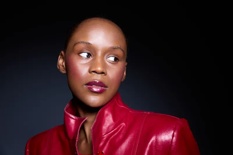 Portrait of a confident young African woman with dark skin wearing a red leather jacket looking sideways. Studio lighting highlights her smooth skin and glossy lips on dark background.