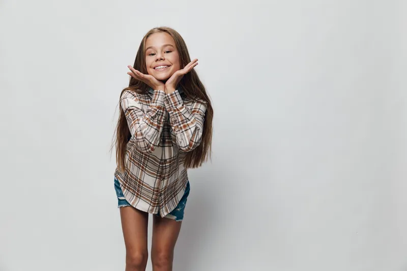 Girl standing with hands near face smiling and looking happy casual outfit on gray background for childhood fun and joy expression lifestyle young person.