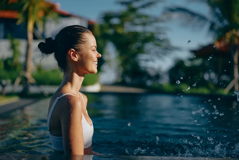 Young woman enjoys relaxing in outdoor swimming pool with eyes closed and smiling face, sunlight illuminates water droplets and tropical background.