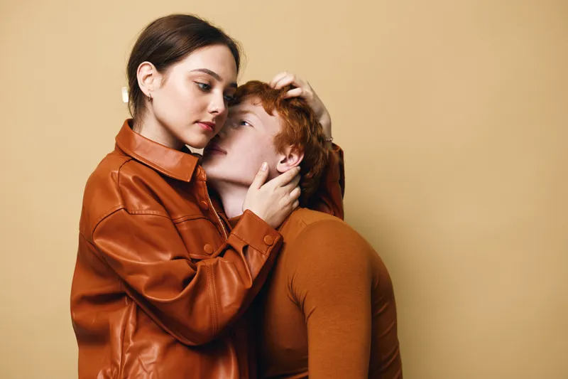 Couple in brown and orange clothing, woman gently holding man’s head with affection, studio portrait with neutral beige background, intimate connection between two young adults.